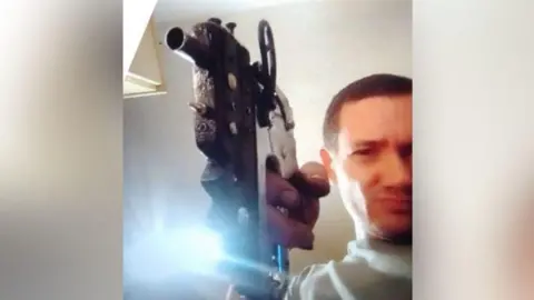 Head and shoulders of a man in his 40s with short brown hair, looking into the camera, holding a converted staple gun. He is pointing it at the camera. 