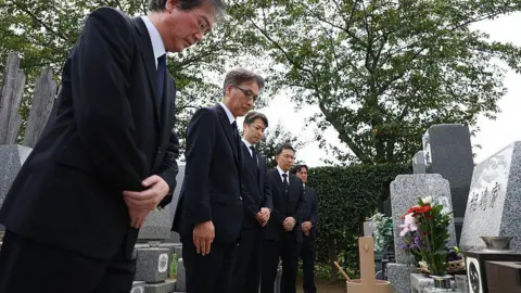 JijiPress via Getty Images Five men wearing dark suits bow before a grave in Japan. The grave is made of grey marble, and has flowers in a vase in front of it