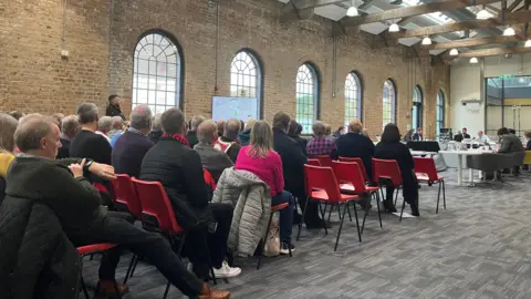 Shariqua Ahmed/BBC Rows of people seated on red chairs in a large room. They are facing committee members, who are seated in an oval-shaped arrangement.
