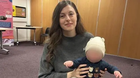 A young woman with long brunette hair wears a grey jumper. She has a smiling baby boy on her knee - he wears a white hat, navy jumper with bears on it and grey leggings.