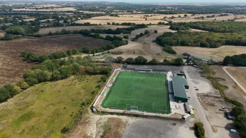 An astroturf football pitch, seen from above. It is surrounded by yellow countryside.