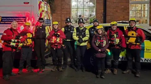 West Mercia Search and Rescue Volunteers from West Mercia Search and Rescue with three police officers. They are standing in front of two emergency response vehicles, a van on the left and a car with green and yellow markings on the right. 