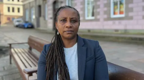A woman called Deputy Beatriz Porée sits on a wooden bench in front of a pink building in St Helier, Jersey. Porée is a black woman with long and thin dreaded hair which goes down past her chest. She is wearing a navy blue blazer with a grey top underneath. She has large silver earrings and a necklace on.