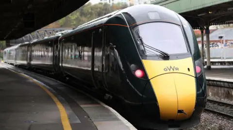 A GWR train pictured at a train station. It has a yellow front with red headlights and a dark green body. 