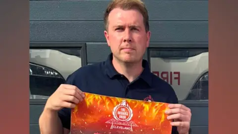 James Sudron is holding up a large poster that has a design depicting orange and red flames. A white logo at the top reads: "Fire Brigades Union Cleveland". He has short brow hair and is wearing a blue polo shirt.