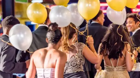 Pictured is a hall full of teenagers in prom dresses and suits. They are holding silver and gold balloons.