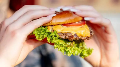 Getty Images A close up of two hands holding a burger. There is a toasted brown bun, moist meat with melted cheese on top. A cheeky tomato slice is peeking out of the top of the bun and an large piece of lettuce at the bottom.