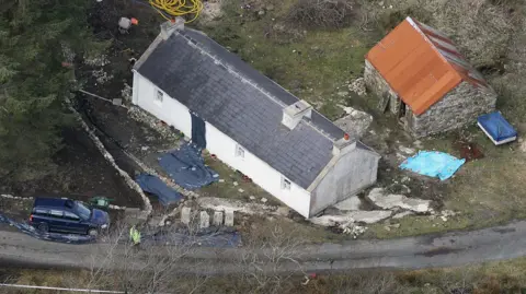 Getty Images An aerial image of a white washed house in a remote location. S shed is behind the house and trees are around it. A police officer is standing nearby.