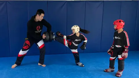 Six-year-old Ivanna performs a high kick during pad work training with her dad and brother. 