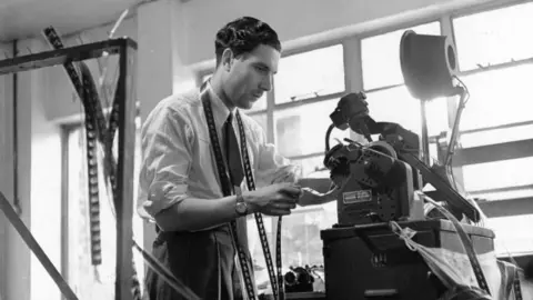 Getty Images A black and white image of a film editor working on a film at Denham Studios. He has machinery in front of him. and he is looking at film. He is wearing a white shirt, has a tie on and trousers. Windows are behind him. 