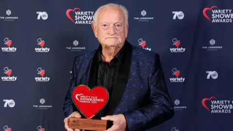 Getty Images Pip Burley poses with a trophy at the Variety club Gold Heart Legacy Award 2025. He wears a navy patterned suit, and black shirt. He has short grey hair and is bald on the top of his head.