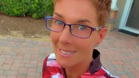 Carmel McConnellogue Carmel McConnellogue has short brown hair and is wearing a Derry City tracksuit top. 
She has purple glasses and her ear is pierced. 