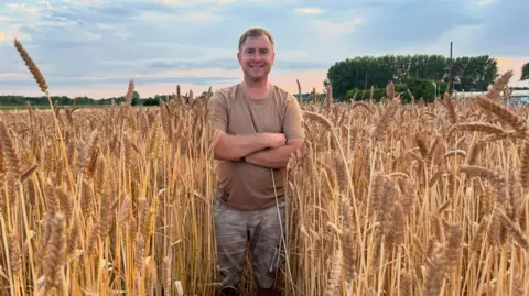 Chris Dodson Chris Dodson smiles at the camera as he stands in a field of straw with his arms crossed across his chest. He is wearing a brown T-shirt and jeans. Behind him is a blue and pink skyline. 