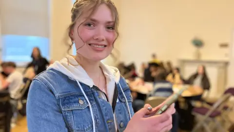 A teenage girl stands holding a phone and smiling at the camera. There are other students sitting at tables, blurred in the background.