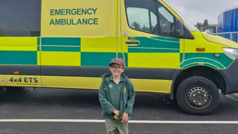 SATH A boy with glasses, a green tshirt and fleece and a red cap standing in front of a green and yellow chequed ambulance