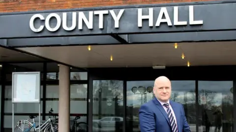Local Democracy Reporting Service Man in navy blue suit standing outside building with the words "COUNTY HALL" In bold white lettering 