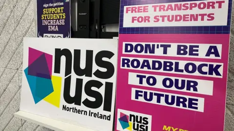 NUS-USI placards leaning against a wall. They are coloured - white, pink, yellow and blue. One says NUS-USI Northern Ireland, another says 'Don't be a roadblock to our students'.