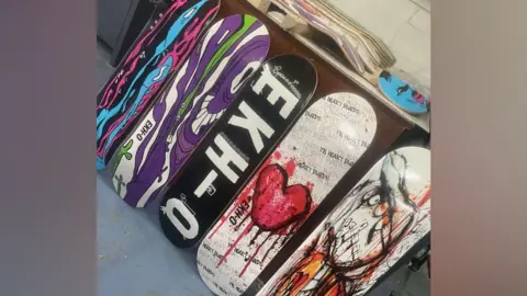Rachael Bassett A collection of skateboards which have been painted on with different artworks rest against a table. 