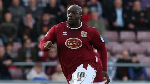 A man in a claret football shirt and white shorts jogging on a football pitch. Spectators can be seen in the background. 