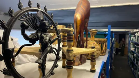 A collection of artefacts on a blue shelf at the museum store. One piece is in the shape of a decorative wheel with a figurine in the centre. Behind it are wooden carved pieces including a vase.