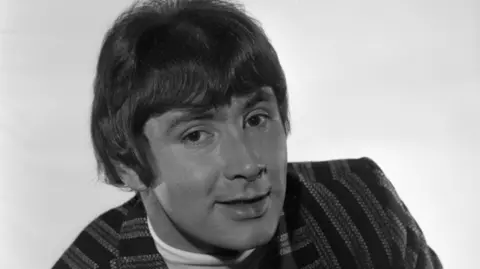 A black and white photograph of Reg Presley half-smiling and wearing a pinstripe suit jacket.