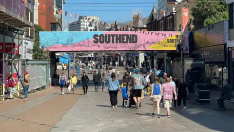 People walking up and down the pedestrianised Southend High Street. There is a large overhead banner saying SOUTHEND on a blue, pink and yellow background