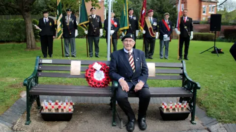 Alan Bray A man wearing clothes including a jacket, a tie and trousers is sitting on a bench next to a wreath and looking at the camera. Eight people are standing up on grass behind him.