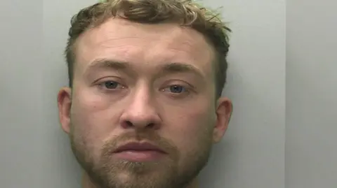 A mugshot of Jamie Hawken. He is staring at the camera. He has facial hair and brown/blonde hair. 