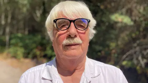 Mike Briggs with white hair, glasses, and a white button-up shirt standing outdoors on a sunny day. The background shows a tree-lined path with softly blurred greenery.