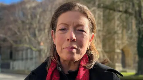 A head and shoulder shot of a woman with dark eyes and shoulder-length brown hair. She is wearing a black coat and a red and black scarf. She is stood in front of Beverley Minster with the background out of focus.