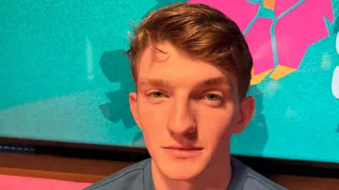 Jamie Jennings is looking into the camera sitting on a red sofa with a State of Us branded wall behind him. The wall is blue/green with a pink and orange outlined NI map. Jamie has short light brown hair and is wearing a blue jumper.