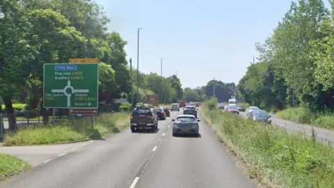 Google Vehicles approacing the Lawnswood roundabout in Leeds, with a large green road sign on the left, indicating the oncoming roundabout