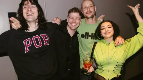 Henry Redcliffe Four people posing for a photo, Charli XCX is wearing a lime green corset hoodie, the other people from left to right are wearing a black jumper with the word POP in red letters, a black shirt and another a green jumper