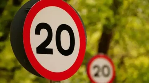 A 20mph road sign stands before greenery, with another 20mph road sign in the distance.