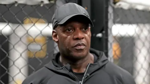 Trevor is a middle aged man wearing a black cap and a black sports top, he is standing in a gym.