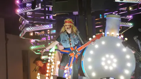 A young person sports a classic rock look, complete with a long, blond, shaggy wig, denim vest, red headband, and a guitar with the Union Flag, while riding a rock-themed float at Bridgwater Carnival