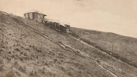 An old postcard in sepia. It shows a lonely building on top of a hill. 