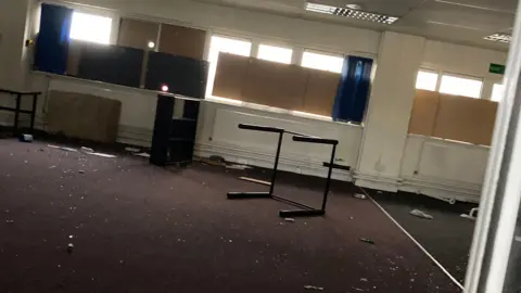 CDDFRS Interior of a derelict office room. Broken furniture is scattered around the room, there are piles of broken glass and the windows are covered in boards