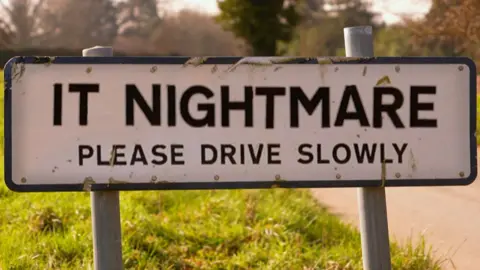 A village road sign displaying "IT NIGHTMARE, PLEASE DRIVE SLOWLY"