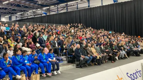 Crowds sit in chairs watching the first games. There are some team members in a blue kit sat on the front row.