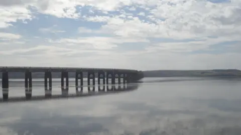 Neil Halley A long bridge stretches across a wide estuary under a partly cloudy sky. The still water mirrors both the bridge and the clouds.