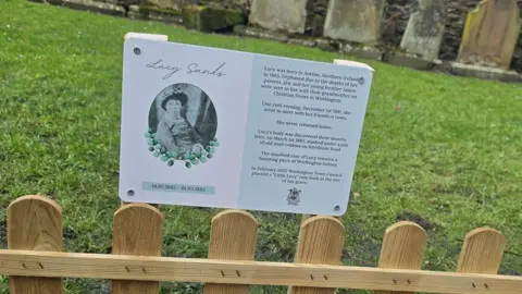 Workington Town Council The small memorial plaque with a black and white picture of Lucy Sands on the left and some words to describe who she was on the right.