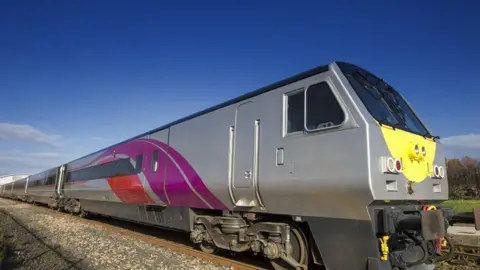 Silver enterprise train, with purple and red stripe branding. On the tracks in the countryside, on a clear sunny day.