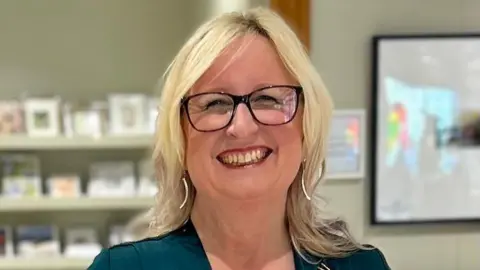 Sharon O'Connell Picture of a smiling blonde lady, with dark glasses in a teal dress. 