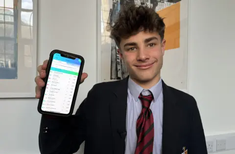 Student in shirt and tie smiling at the camera. He's holding up his phone to the camera and is showing a green and blue league table.