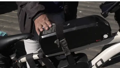 A black battery strapped on to the white frame of a normal push bike to make it an e-bike