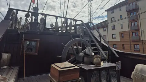 An elevated wooden deck area with extensive rigging lines stretching upward and outward, and a large traditional ship's wheel positioned near the centre.
