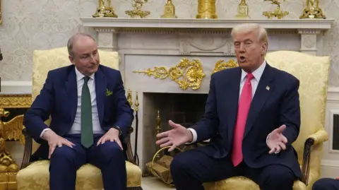 PA Media Micheál Martin (left) and Donald Trump (right) sit on two gold-coloured chairs in the Oval office. Mr Trump is talking while Mr Martin listens. There is a large marble fireplace in the background with a number of gold decorations.
