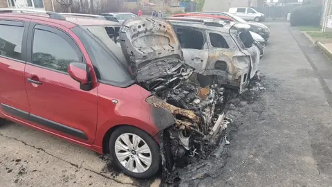 BBC Two cars in a row of parked cars are severely burnt. The nearest one, which is red, has its bonnet open and the engine compartment is a mass of grey, tangled, burnt metal and charred remains. Beside it is a white car which is also burnt but is damaged less.