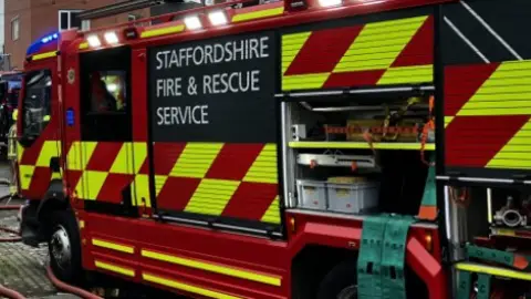 BBC The side of a red fire engine, some of the compartments are open, and pipes  trail on the ground. Staffordshire Fire and Rescue Service is written on the side of the vehicle